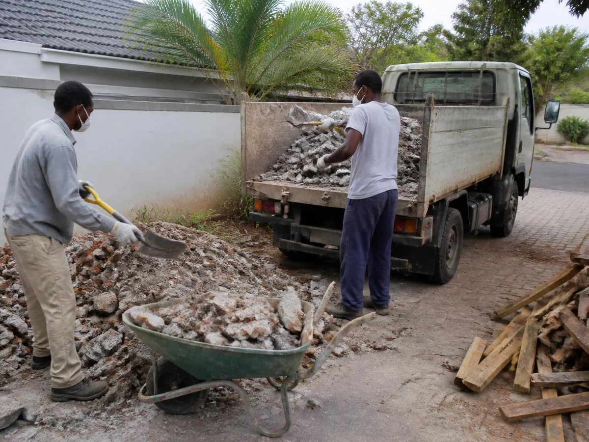 Two workers in gloves and dust masks shovelling broken brick and concrete from a demolition heap into a small tipper truck on a Cape Town driveway, with timber kept in a separate pile