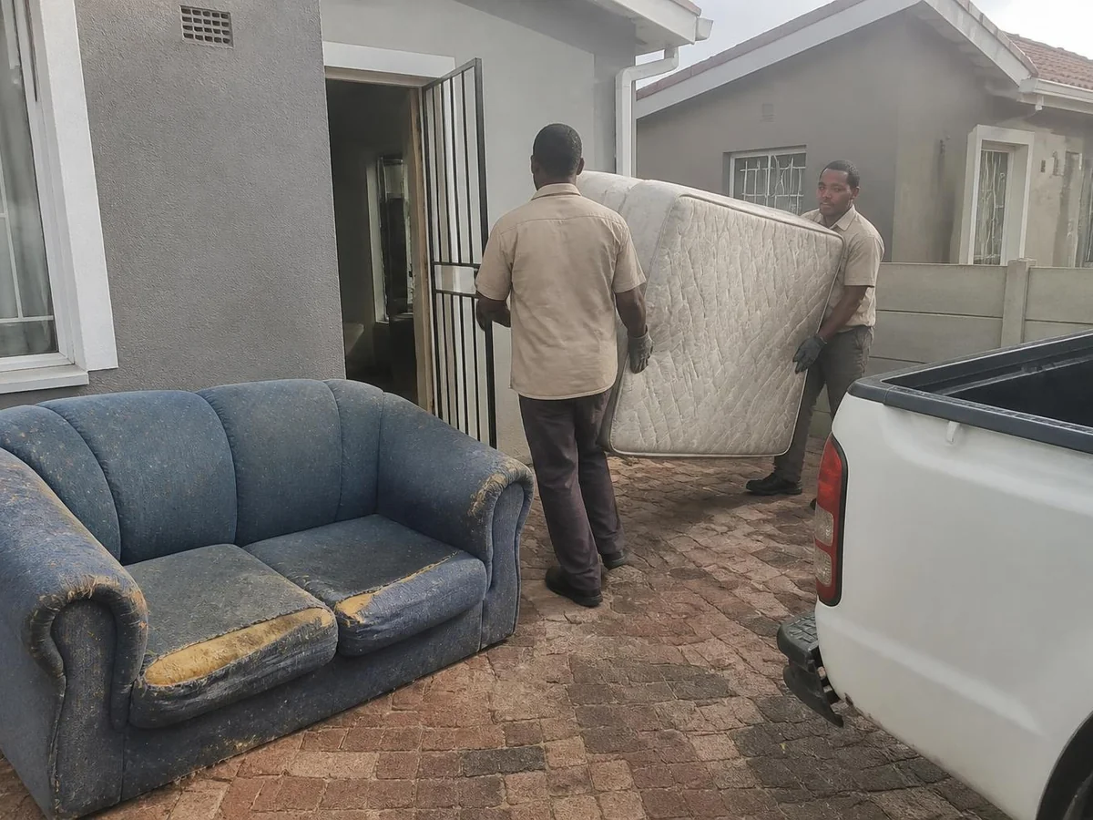 Two workers carrying an old double mattress out through the front doorway of a Cape Town house to a bakkie, with a worn couch already standing on the paving